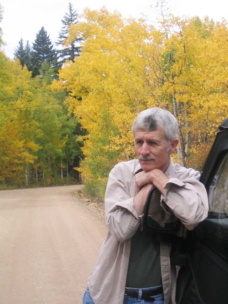 George Drew leans against the side of a truck, a dirt road lined with fall-colored trees behind him